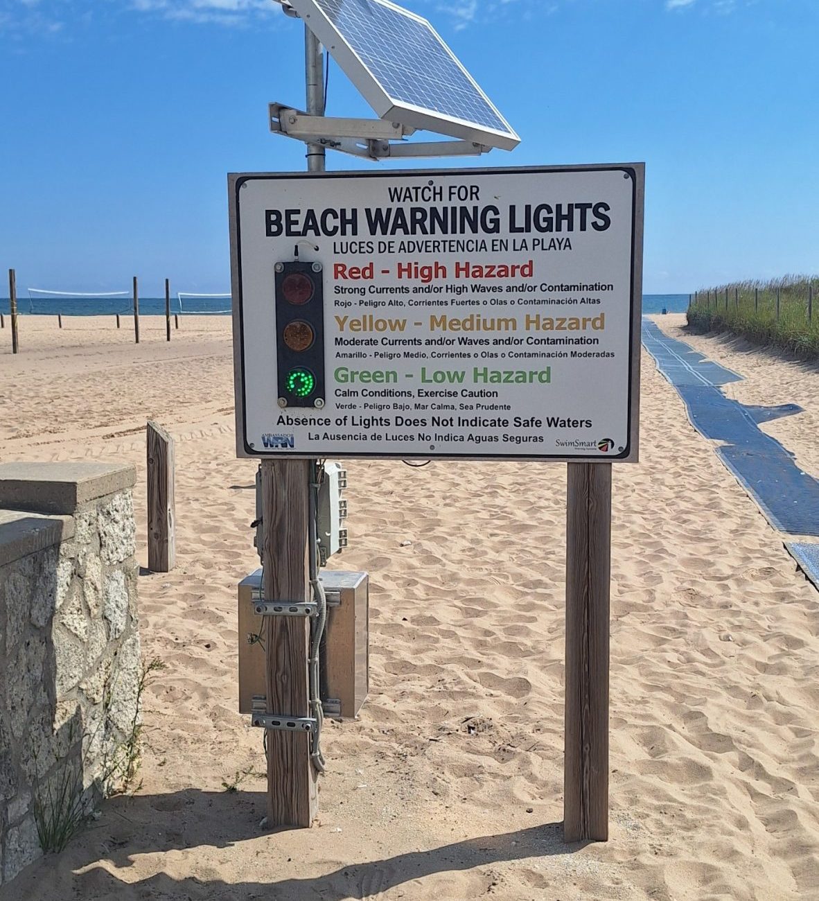 Beach warning sign with color-coded hazard levels (red, yellow, green) powered by a solar panel on a sandy beach.