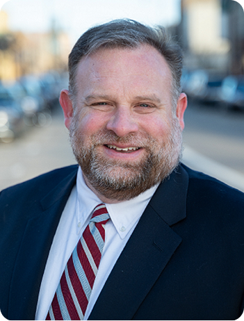 Headshot of a smiling man wearing a suit and tie, outdoors with blurred background.