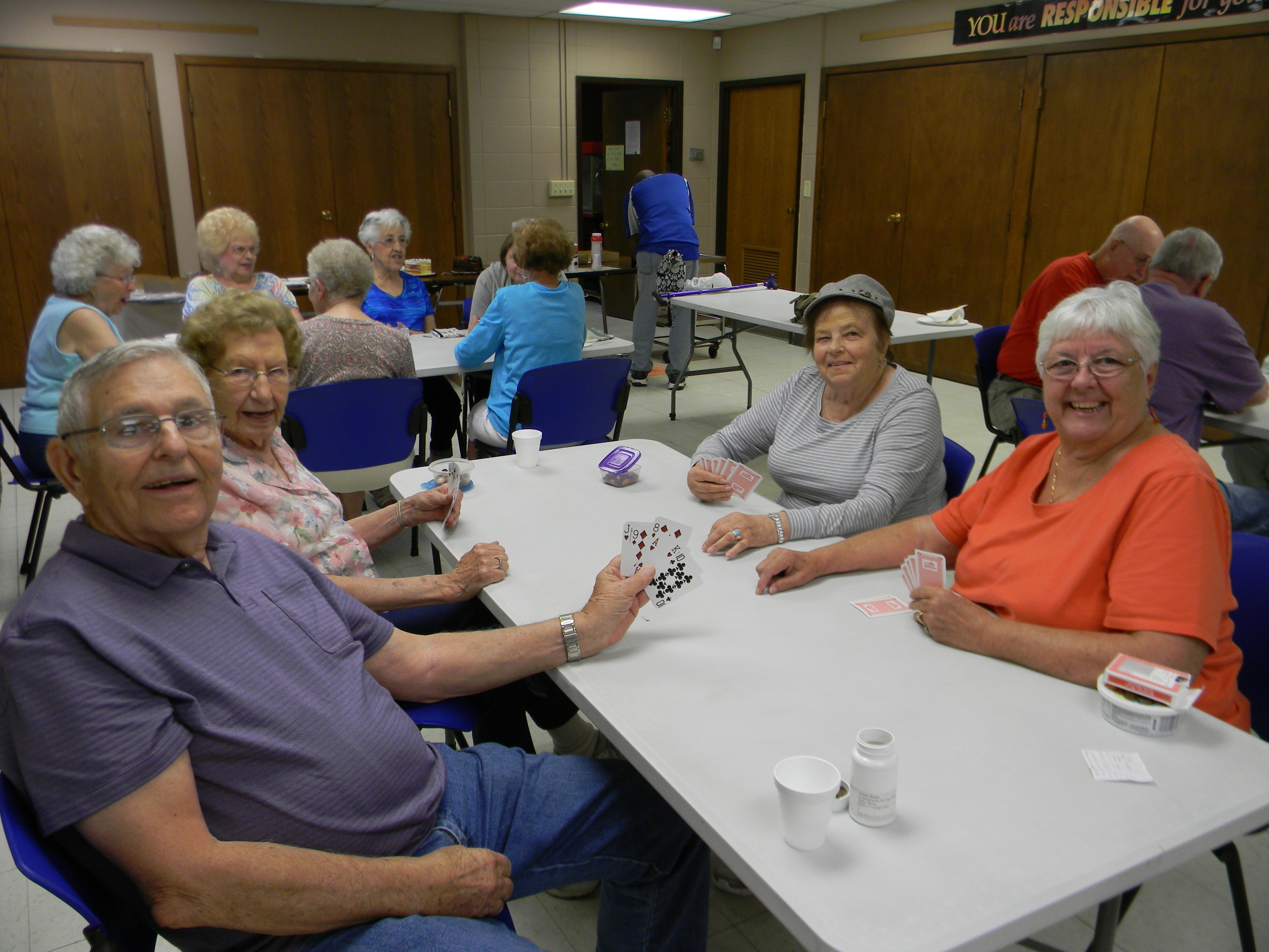 Group of seniors playing cards at tables in a community center, smiling and engaged in the game.