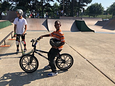 Young boy on BMX bike in skatepark, holding helmet, with another boy on scooter in background.