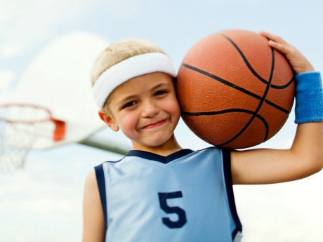 Young boy in basketball uniform with ball on shoulder, smiling. Basketball hoop visible in background.
