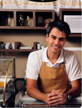 Smiling barista in apron leans on counter in a cafe, shelves with glassware and coffee supplies in background.