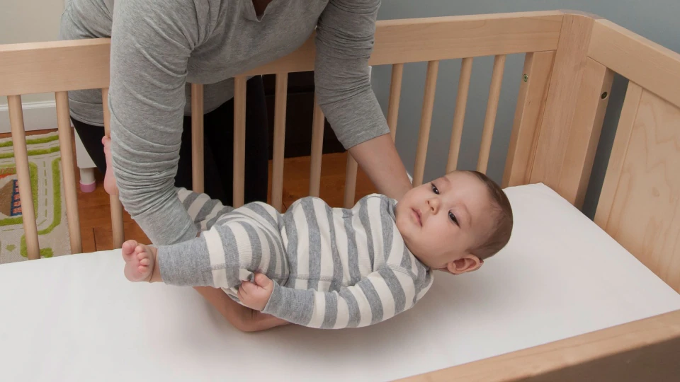 Adult placing a baby in a striped outfit into a wooden crib with a white mattress.