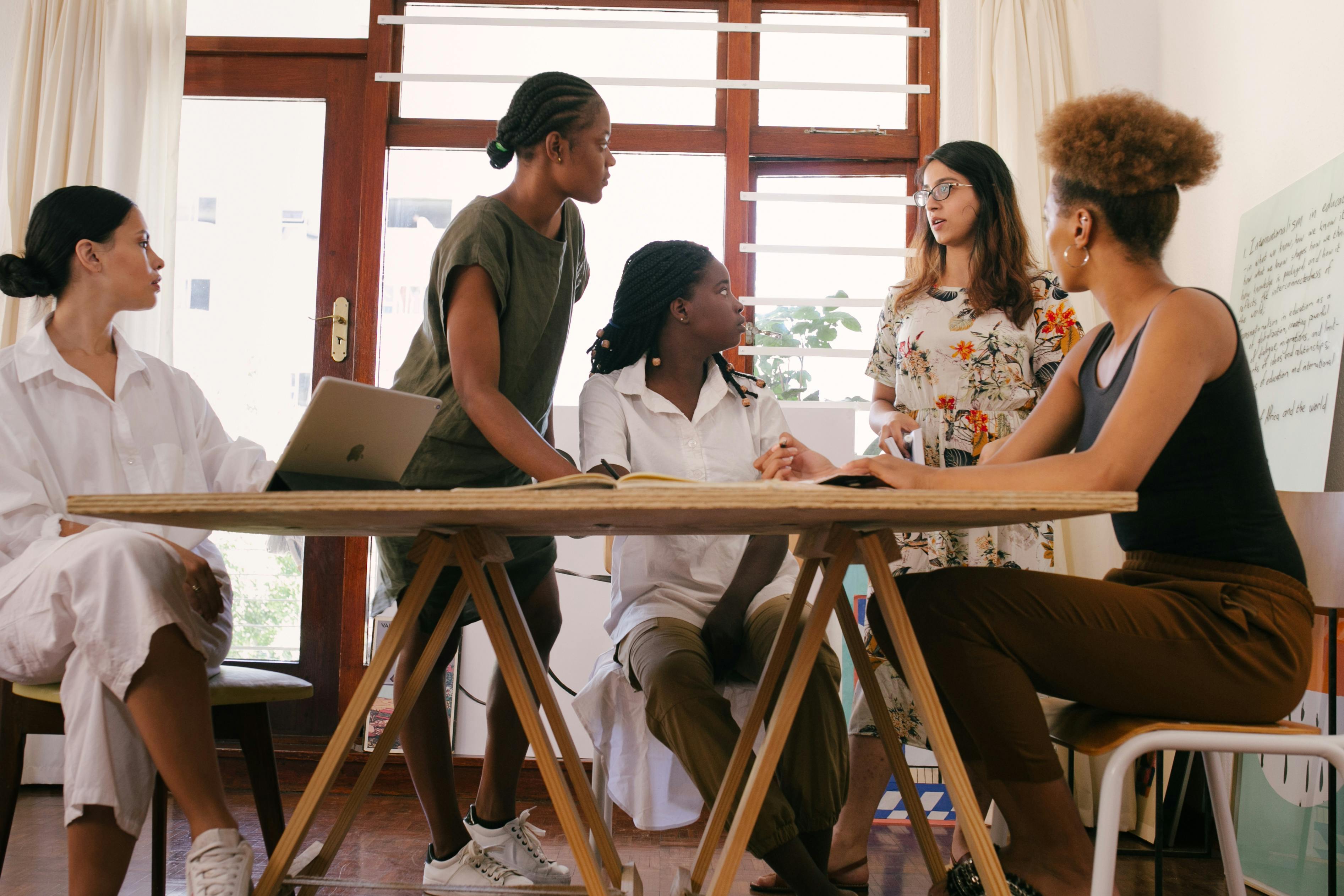 Diverse group of women collaborating around a table with a laptop and notes in a bright, modern office space.