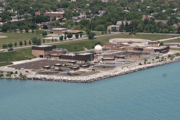 Aerial view of a wastewater treatment plant on a coastline, with tanks, buildings, and a spherical structure.