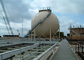 Spherical storage tank with access stairs at an industrial facility, with cranes visible in the background.