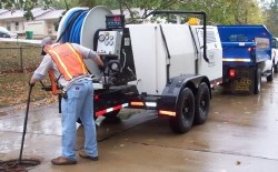 Worker using a sewer jetter to clear a manhole, with truck and trailer equipment nearby.