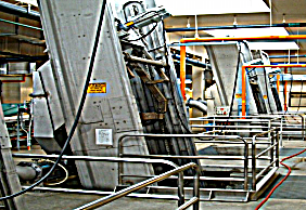Industrial machinery with metal framework and safety railings in a factory setting. Overhead pipes are visible.