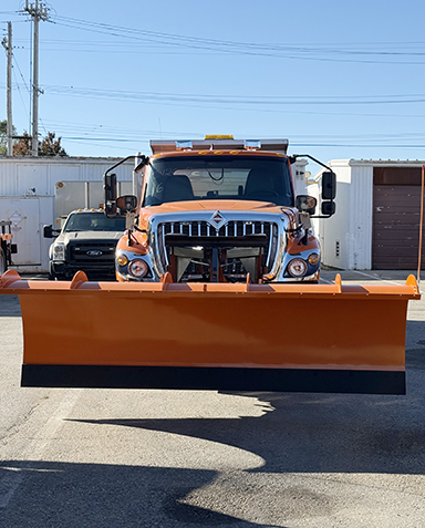 Orange snowplow truck parked on pavement, front view. White building and truck in background under a clear blue sky.