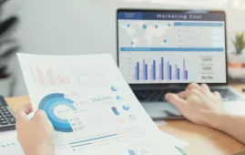 Hands holding marketing data report with laptop showing marketing goal charts in background.