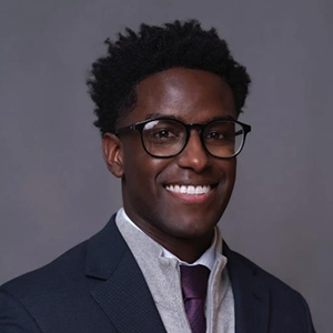 Professional headshot of a smiling Black man wearing glasses, a suit jacket, and a tie against a gray background.