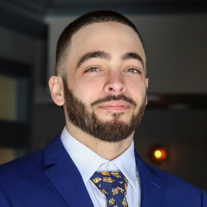 Headshot of a man with a beard, wearing a blue suit and patterned tie, looking directly at the camera.