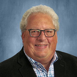 Headshot of smiling man with glasses, gray hair, wearing a blue checkered shirt and black blazer against a blue background.