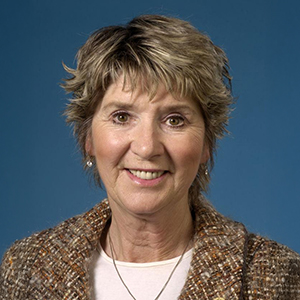 Headshot of a smiling woman with short blonde hair wearing a patterned jacket against a blue background.