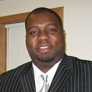 Professional headshot of a Black man in a pinstripe suit, smiling slightly. He has short hair and a goatee.