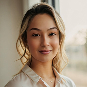 Portrait of a young woman with blonde hair, wearing a white shirt, standing near a window.