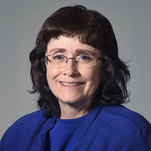 Headshot of a woman with glasses and a blue shirt, smiling slightly against a gray background.