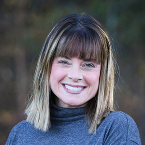 Headshot of a smiling woman with bangs and a gray turtleneck sweater against a blurred outdoor background.