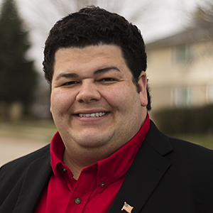 Headshot of a smiling man in a red shirt and black jacket, wearing an American flag pin.