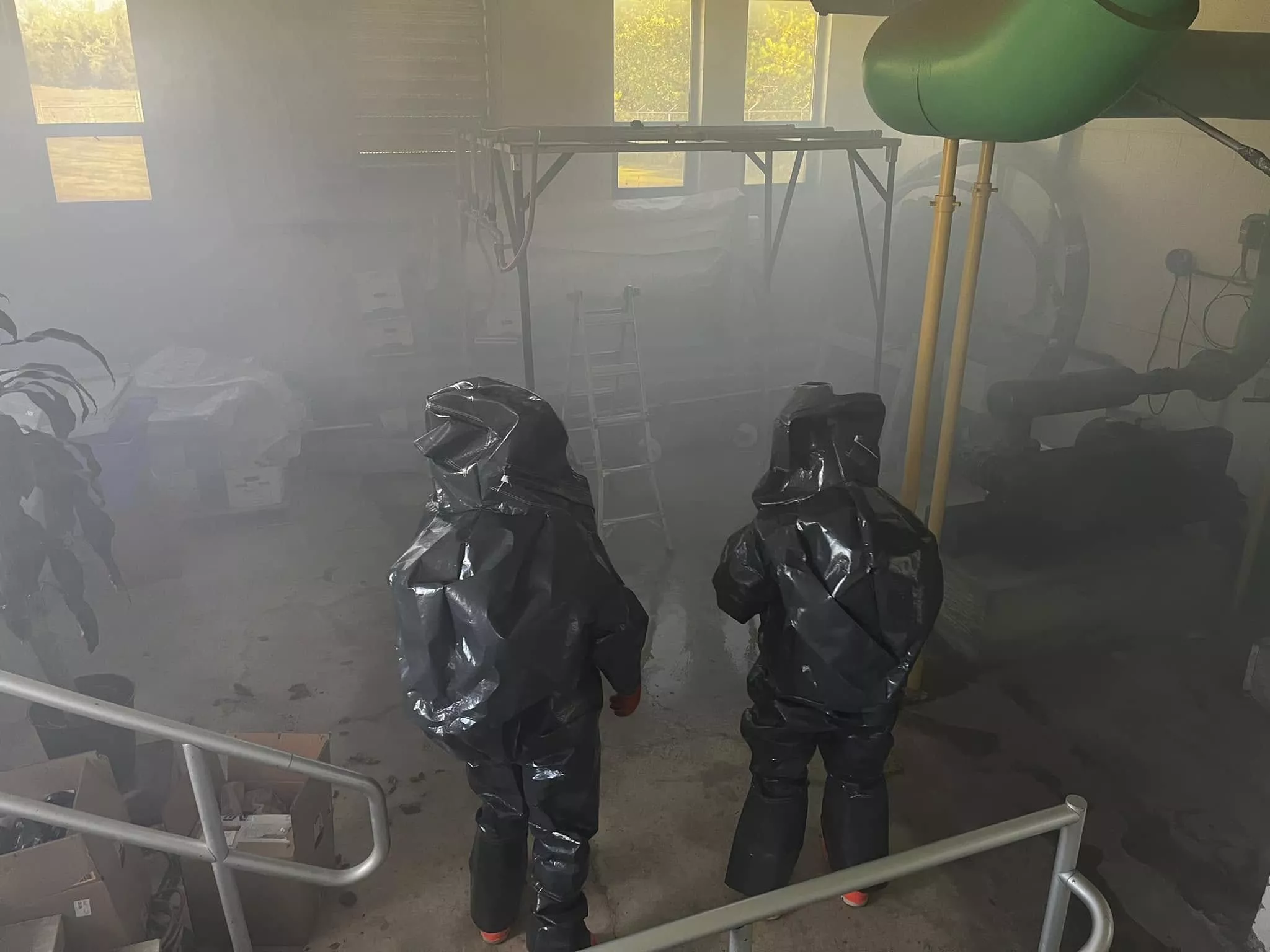 Two people in protective suits stand in a smoky room with equipment and a window in the background.