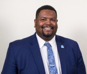 Professional headshot of a smiling man wearing a blue suit and patterned tie against a neutral background.
