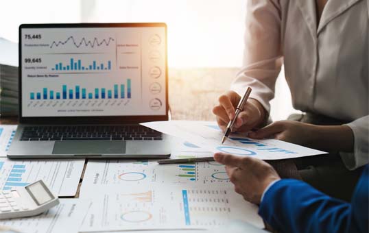 Two people analyzing financial data on a laptop and printed reports, using a pen to highlight information.
