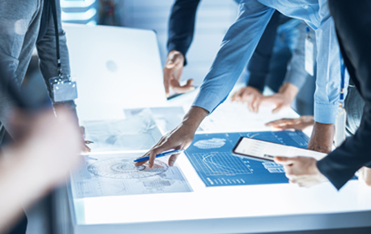 Team collaborating on technical plans with laptop, tablet, and blueprints on a brightly lit table.