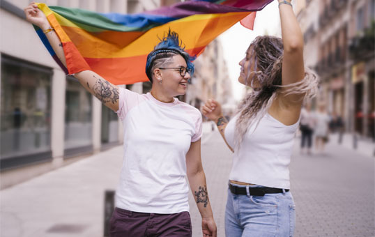 Two women holding a rainbow flag, smiling at each other in a city street. Celebrating LGBTQ+ pride.