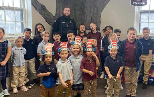 Group of children wearing Cat in the Hat hats pose with a police officer in front of a tree mural.