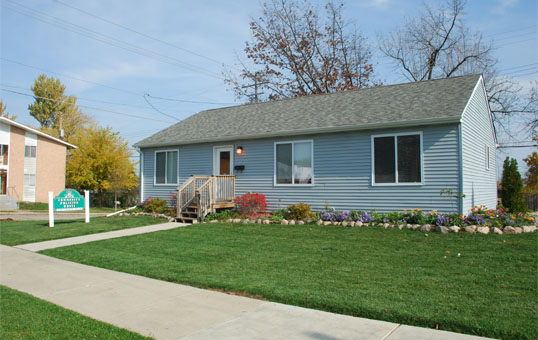 Light blue single-story house with a green lawn, sidewalk, and sign in front on a sunny day.