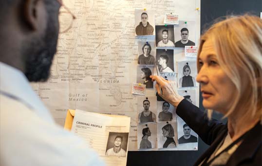 Two detectives review suspect photos pinned to a map during a criminal investigation.