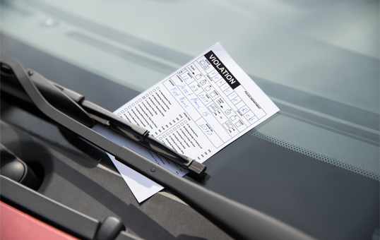 Parking violation ticket under windshield wiper on a car. The ticket is white with black text and boxes.