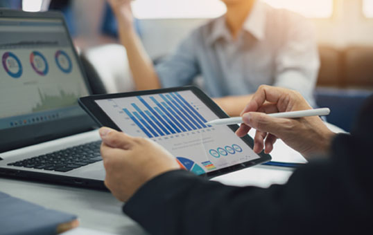 Person analyzing business data charts on a tablet, with laptop and colleagues in the background.
