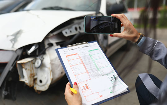 Person documents car accident damage with phone and insurance form on clipboard for claim assessment.