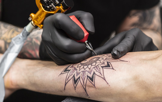 Tattoo artist in black gloves inking a mandala design on a person's arm with a tattoo machine.