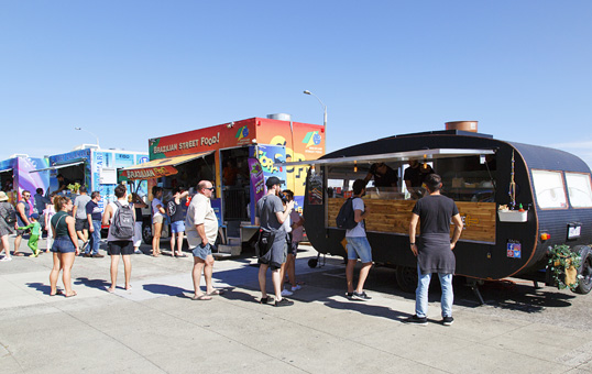 People queue at colorful food trucks on a sunny day, ordering food. Various truck designs are visible.