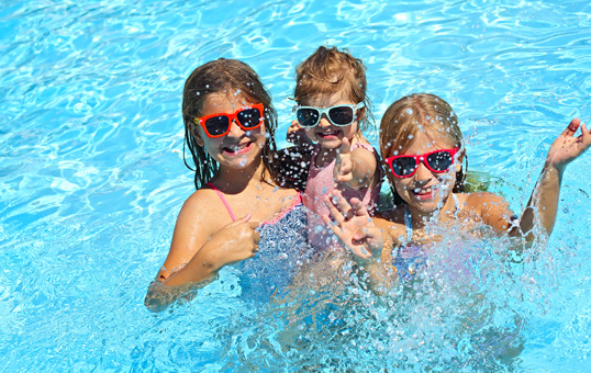 Three smiling girls wearing sunglasses playing in a swimming pool with splashing water.