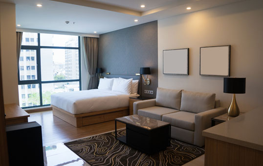 Hotel suite with king bed, sofa, coffee table, and city view through large window. Modern interior design.