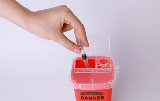 Hand disposing of used syringe into a red sharps container with a clear lid, against a white background.
