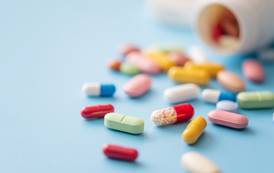 Pills of various colors and shapes scattered on a blue surface, with a pill bottle in the background.