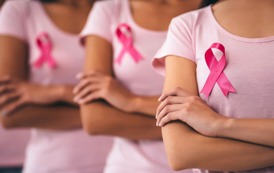 Three women in pink shirts with pink ribbons, supporting breast cancer awareness, arms crossed.