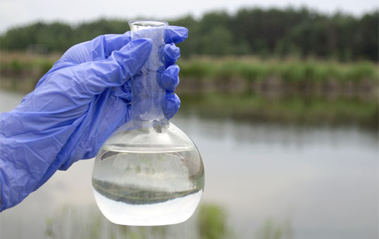 Gloved hand holding a flask of clear water sample, with a lake and trees in the background.