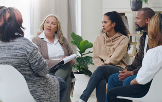 Group therapy session with diverse participants seated in a circle, listening to a facilitator with a clipboard.