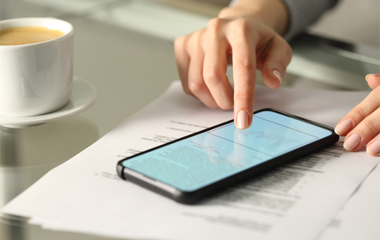 Hand using smartphone on top of documents, with coffee cup visible. Mobile technology for business.