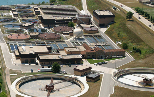 Aerial view of a wastewater treatment plant with circular tanks and rectangular settling basins near a body of water.