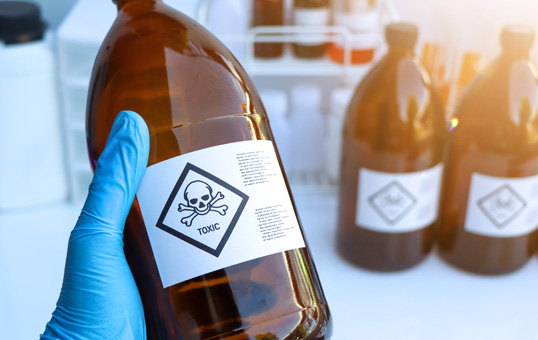 Hand in blue glove holds amber bottle with skull and crossbones 'TOXIC' label. Other bottles blurred in background.