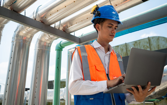 Engineer in hard hat and safety vest using laptop near industrial pipes, performing inspection or maintenance.