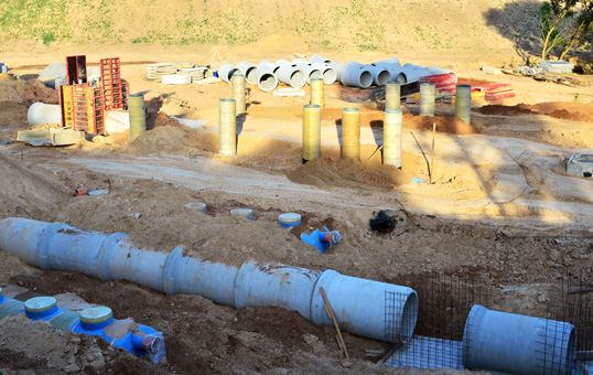 Construction site with large concrete pipes, pillars, and other building materials. Ground is mostly dirt.