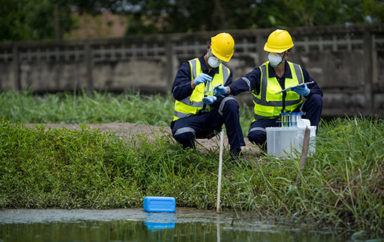 Two environmental scientists in PPE taking water samples from a pond near a concrete wall.