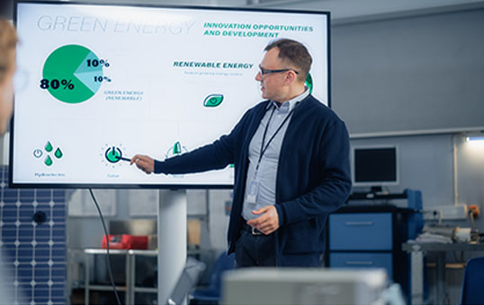 Man presenting green energy data on a large screen, highlighting renewable energy sources with a pointer.